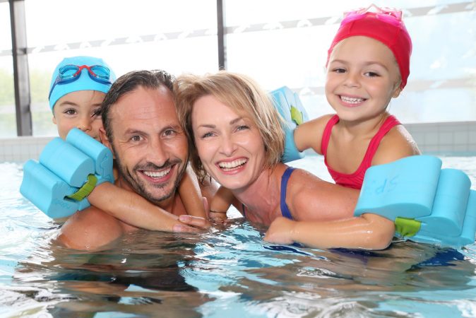 A family in a swimming pool, with children sat on their parents backs and wearing arm bands